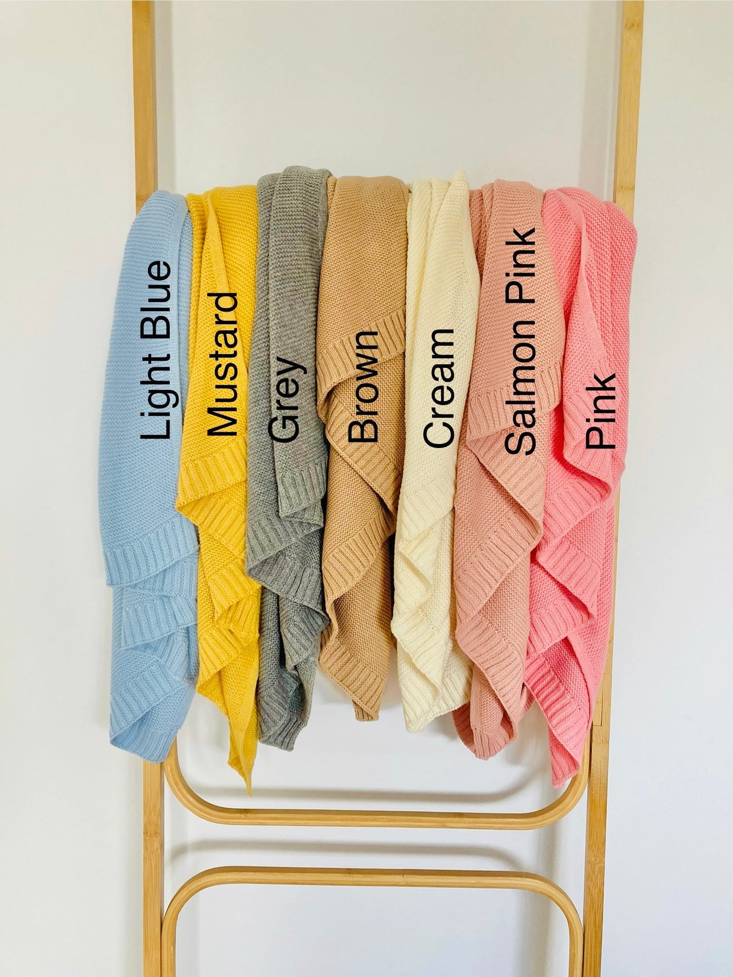 Cotton knit baby blankets with ribbed edge. Light blue baby blanket, mustard baby blanket, grey baby blanket, brown baby blanket, cream baby blanket, salmon pink baby blanket and pink baby blanket draped on a rack. Bespoke Munchkin