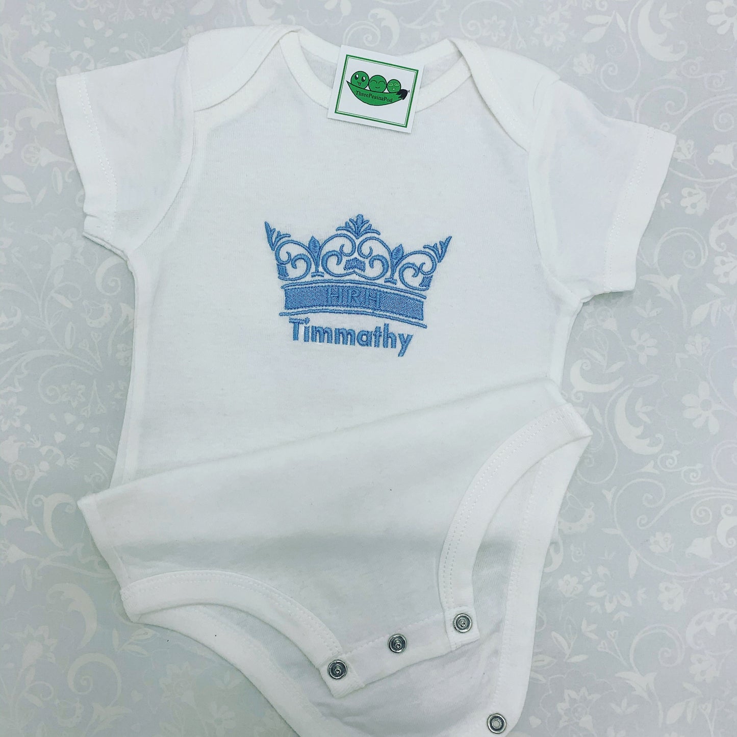 Personalised baby bodysuit with embroidered crown. The crown has the letters HRH on the band of the crown. Baby's name is embroidered underneath the crown. Embroidery in light blue thread.