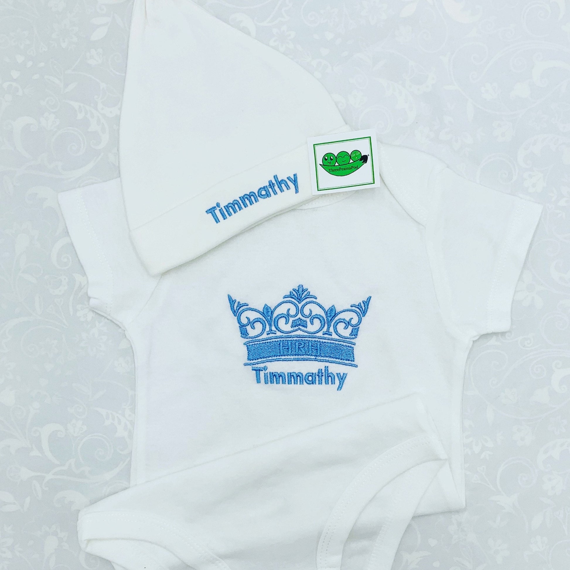 Personalised baby bodysuit with embroidered crown. The crown has the letters HRH on the band of the crown. Baby's name is embroidered underneath the crown and Baby beanie is also customised with baby's name in the same thread colour, light blue.